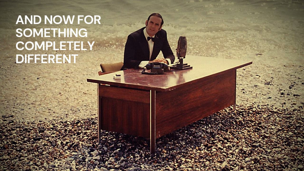 Title card from Monty Python's And Now for Something Completely Different: a man in a dinner jacket sits at a wooden desk on a pebble beach with a vintage microphone and telephone, waves breaking behind him.